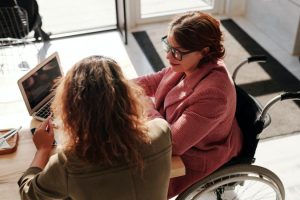 A woman who is a wheelchair user seated at a desk with another woman and an open laptop in an office setting