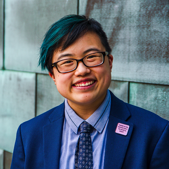 Lydia smiles and tilts their head slightly to the side, looking confidently at the camera. They are a young-ish East Asian person with a streak of teal in their short black hair, wearing glasses, a cobalt blue jacket and navy tie, with a blue copper wall behind them. Photo by Sarah Tundermann.